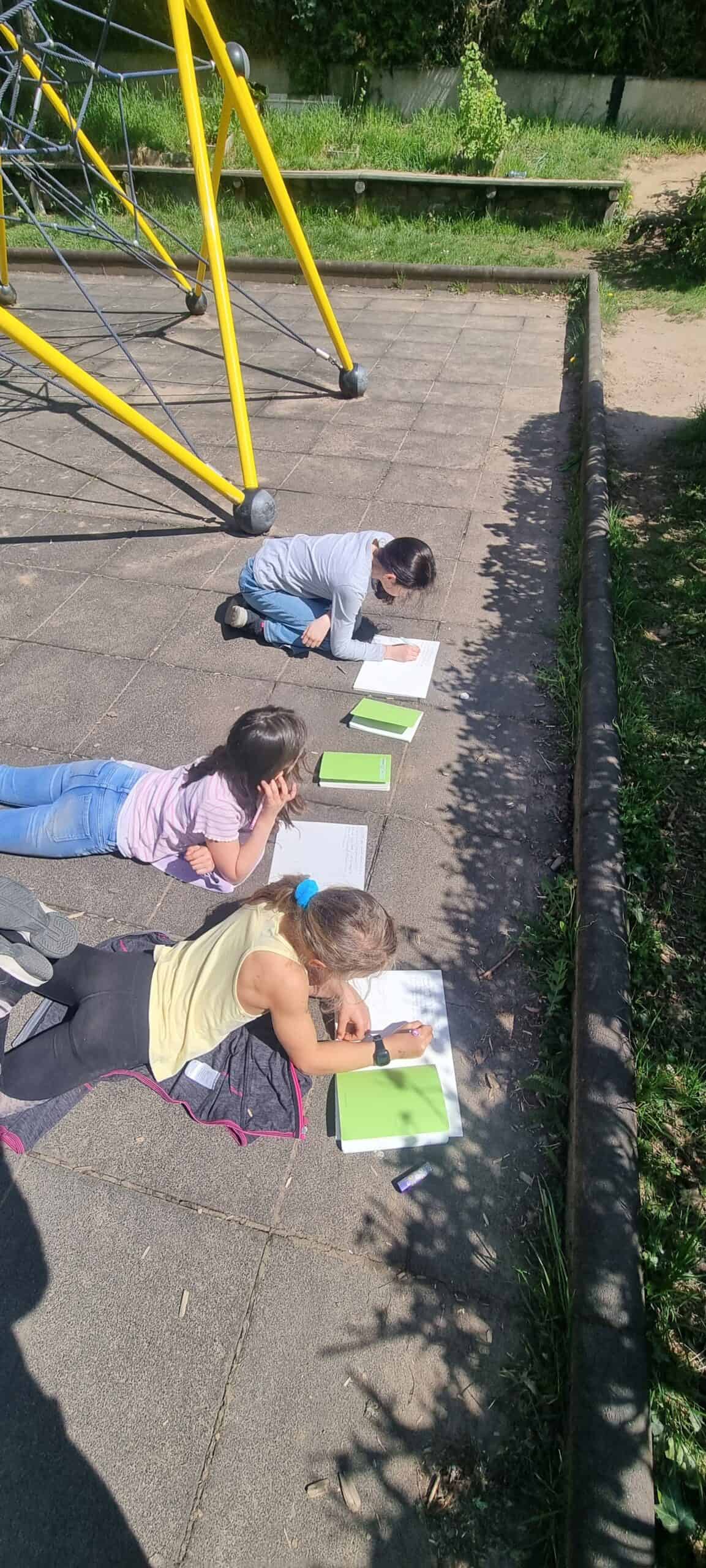 Drei Kinder liegen draußen auf dem Boden. Vor ihnen liegen Zettel und Schreibmaterial.
