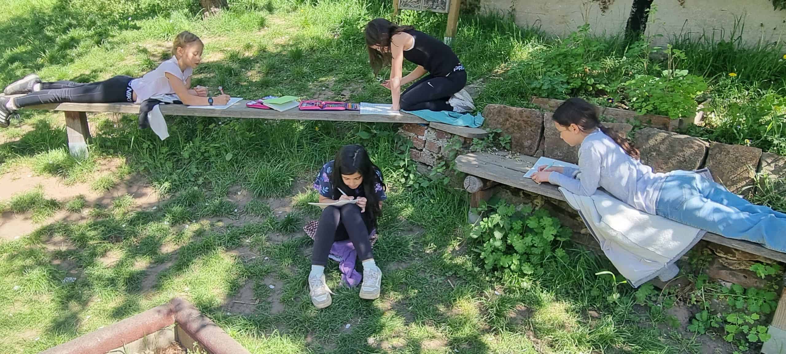 Kinder, die etwas lesen. Ein Kind sitzt auf der Wiese, ein Kind hockt auf einer Bank und ein anderes Kind liegt auf einer Bank.