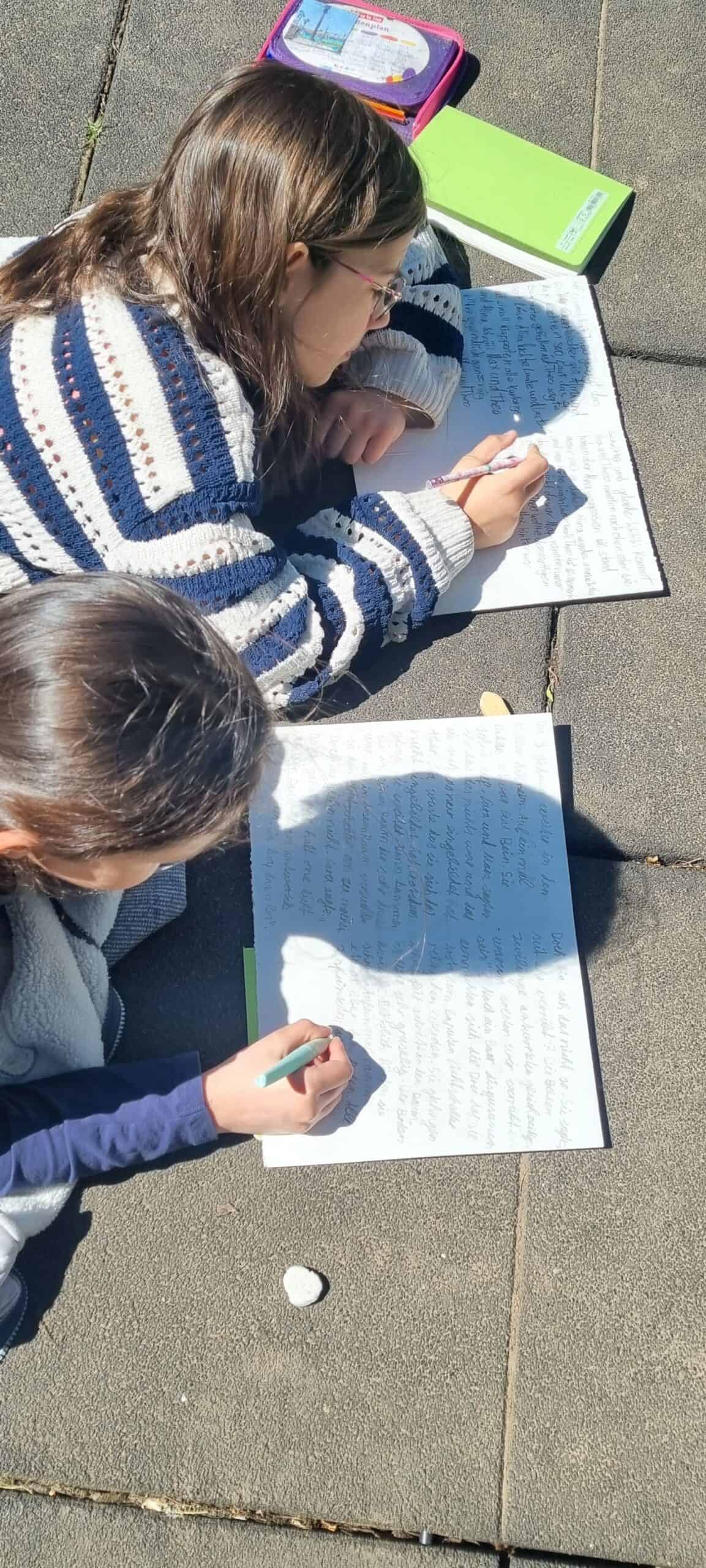 Zwei Kinder aus der Vogelperspektive fotografiert, die auf Betonboden liegen und etwas auf Papier schreiben.